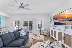Ein Wohnzimmer mit grauem Sofa, einem runden Couchtisch, Deckenventilator, einem großen Fenster mit Sonnenuntergangsblick und einer weißen Kommode mit Fischornamenten.