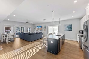 Eine moderne Küche mit einer zentralen Kochinsel, einem Essbereich mit weißem Tisch und Stühlen, einem grauen Sofa und einem großen Fenster mit Ausblick.