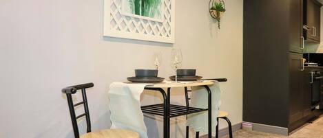 A dining area with a table set for two, chairs, a wall-mounted plant, and a kitchen in the background.