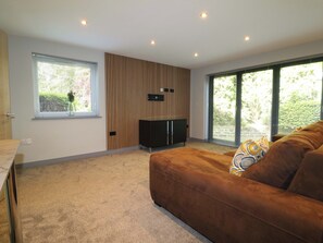 A modern living room with a sofa, a TV, a wooden panel wall, and large windows. - Elmcroft (Nottingham)