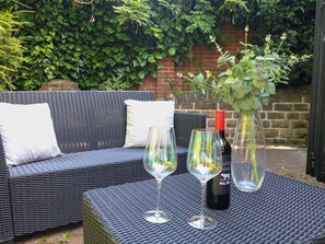 A wicker sofa with cushions, two wine glasses, a wine bottle, and a vase with green foliage. - Elmcroft (Nottingham)