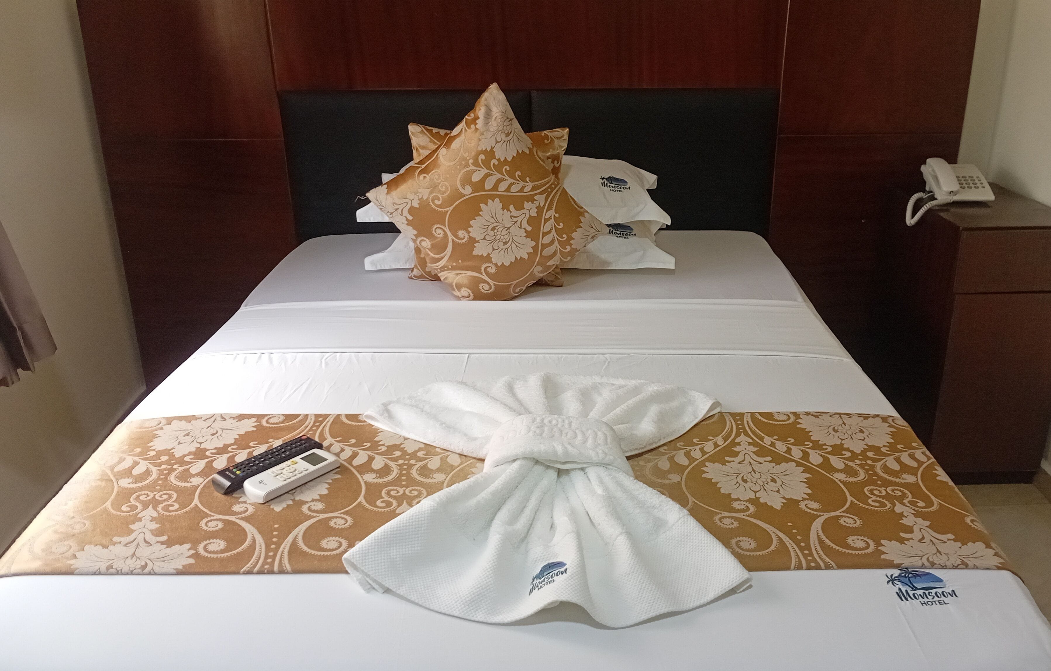 Quarto de hotel com cama bem arrumada, uma almofada, uma toalha dobrada, um controle remoto e um telefone na mesa de cabeceira.