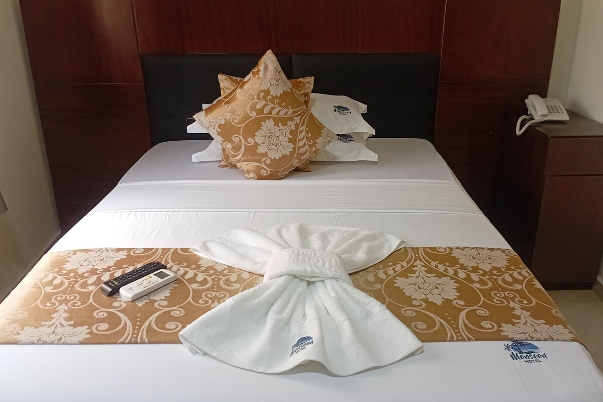 A hotel room with a neatly made bed, a pillow, a folded towel, a remote control, and a phone on the nightstand.