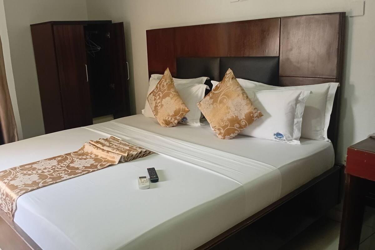 A hotel room with a bed, a wooden headboard, a brown patterned throw, and a remote control on the bed.