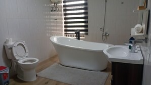 A bathroom with a toilet, bathtub, sink, and a window with blinds. - Monsoon Hotel Ltd (Kumasi)