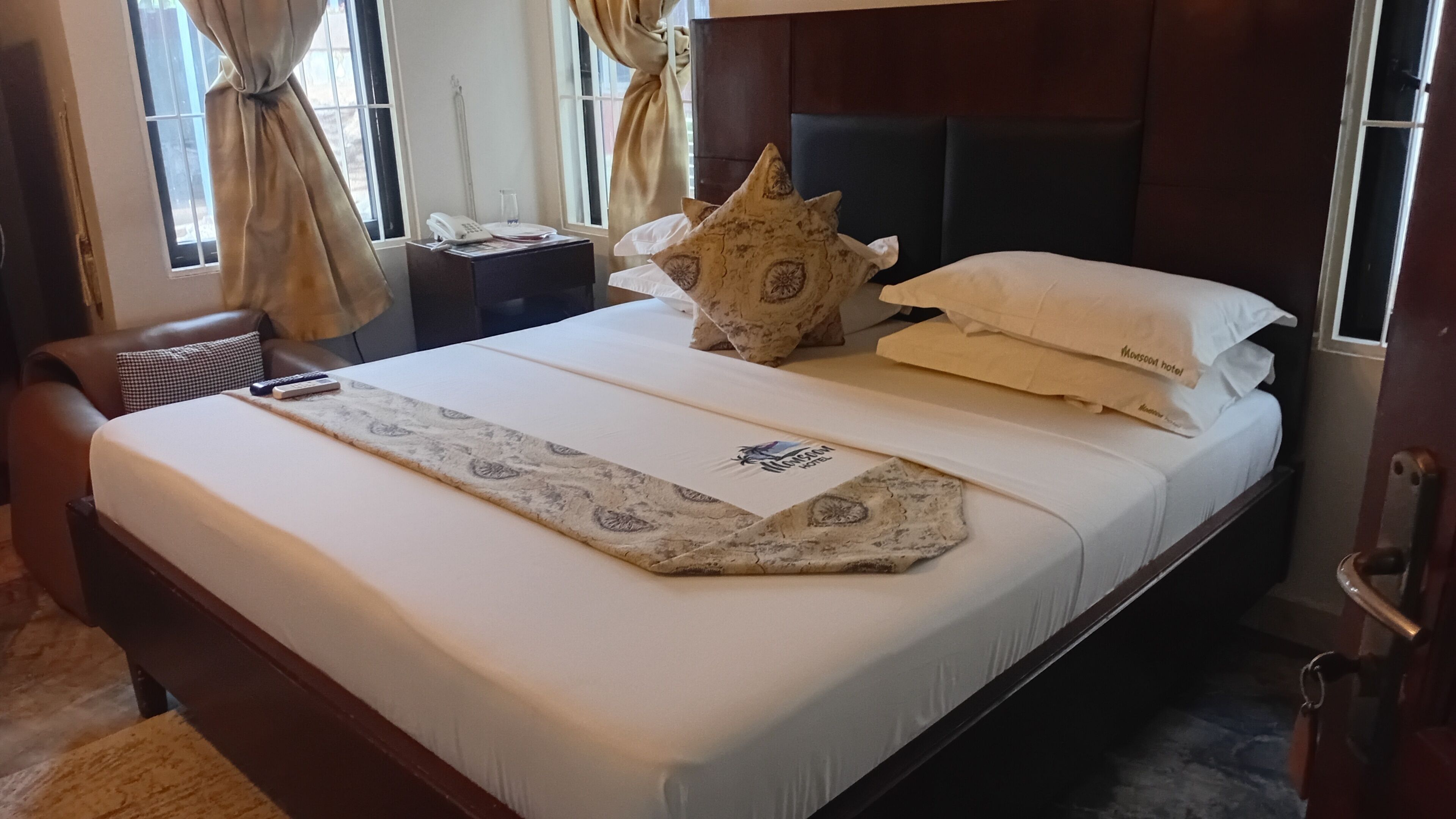 A hotel room with a bed, a bedside table with a telephone, a decorative pillow, and a window with curtains.