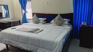 A hotel room with a bed, pillows, a bedside table, a mirror, and curtains. - Monsoon Hotel Ltd (Kumasi)