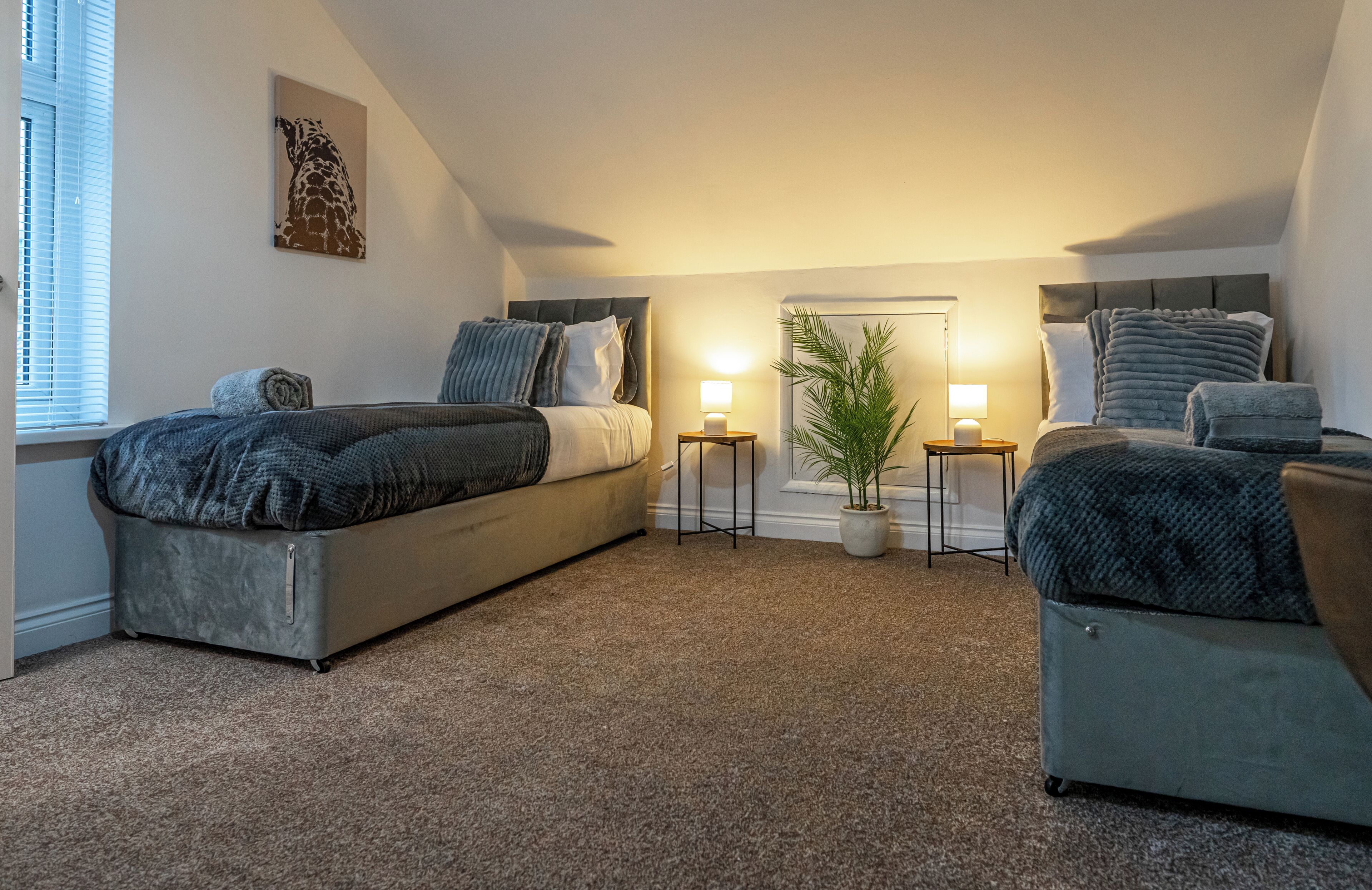A bedroom with a large bed, two bedside tables with lamps, a potted plant, and a giraffe artwork on the wall.