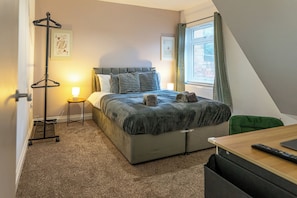 A bedroom with a bed, a bedside table, a lamp, a coat rack, and a window with curtains. - Shrubberies Suite (Derby)
