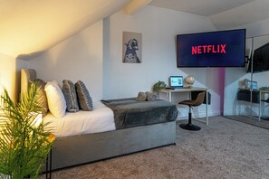 Ein modernes Schlafzimmer mit einem Bett, einem Schreibtisch, einem Stuhl, einem Fernseher, der 'NETFLIX' zeigt, und einem gerahmten Bild an der Wand.