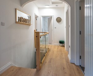 A hallway with a wooden staircase, a mirror with 'HOME' on it, a clock on the wall, and a green stool. - Shrubberies Suite (Derby)