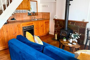 A cozy living area with a blue sofa, wooden cabinets, a stone fireplace, and a kitchen with a sink and stove. - Farr Cottage | Cornish Charm & Unexpected Extras | Pool & Gym Access | Parking (St Ives)