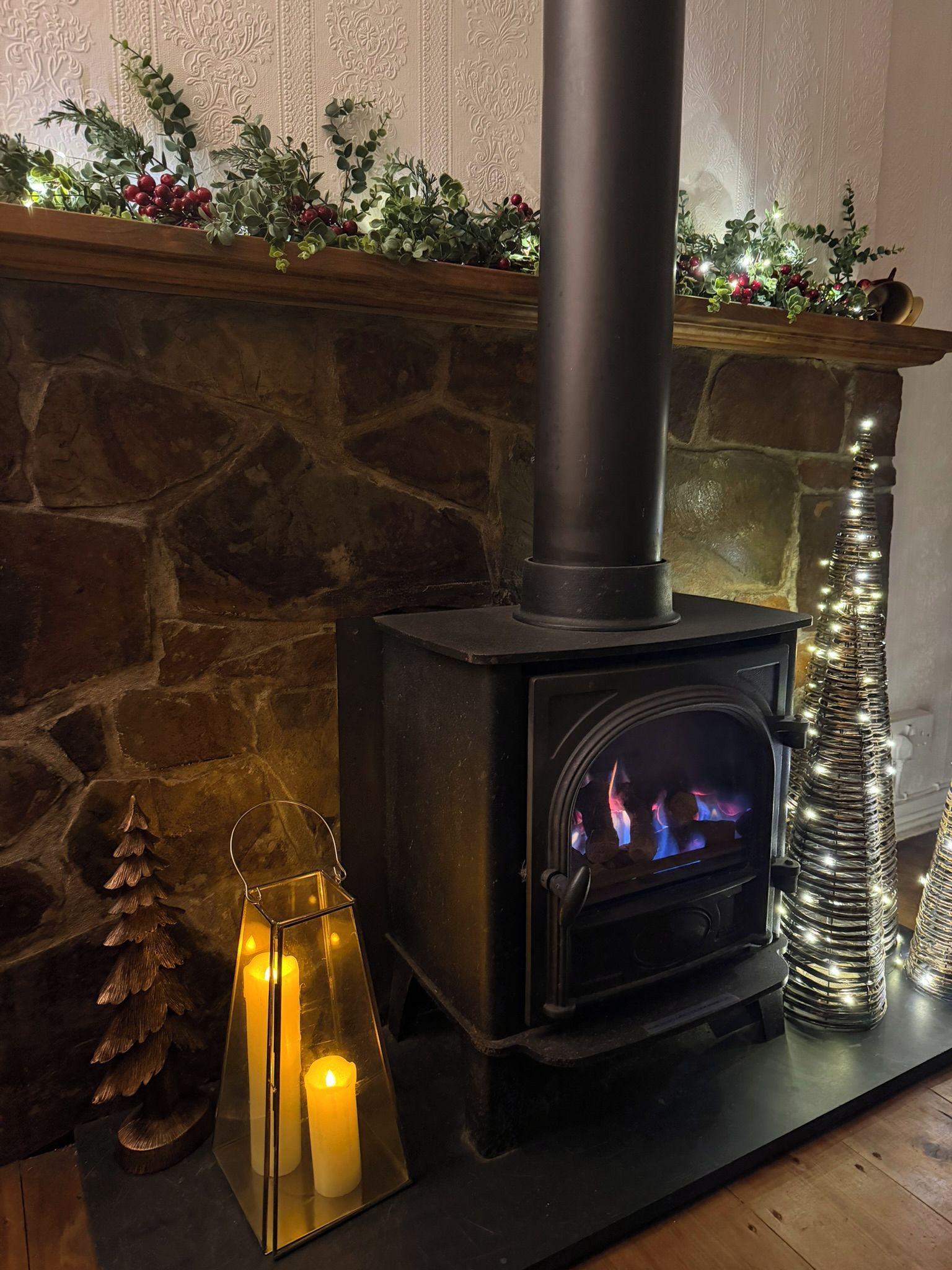 A wood-burning stove with a chimney, a decorative garland above it, and a lit candle in a lantern on the mantel.