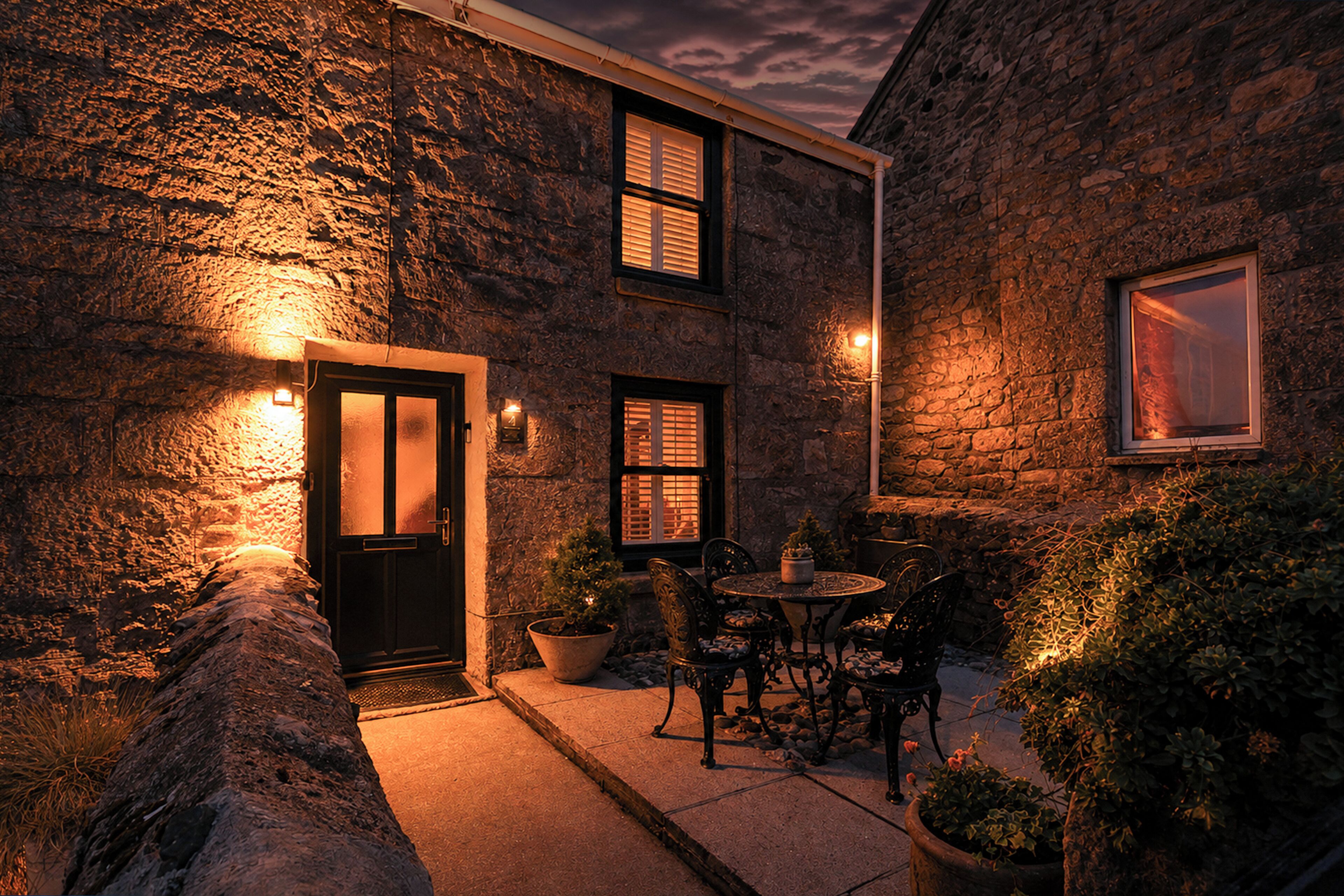 A stone building with a black door, two windows with shutters, a small patio with a table and chairs, and potted plants.