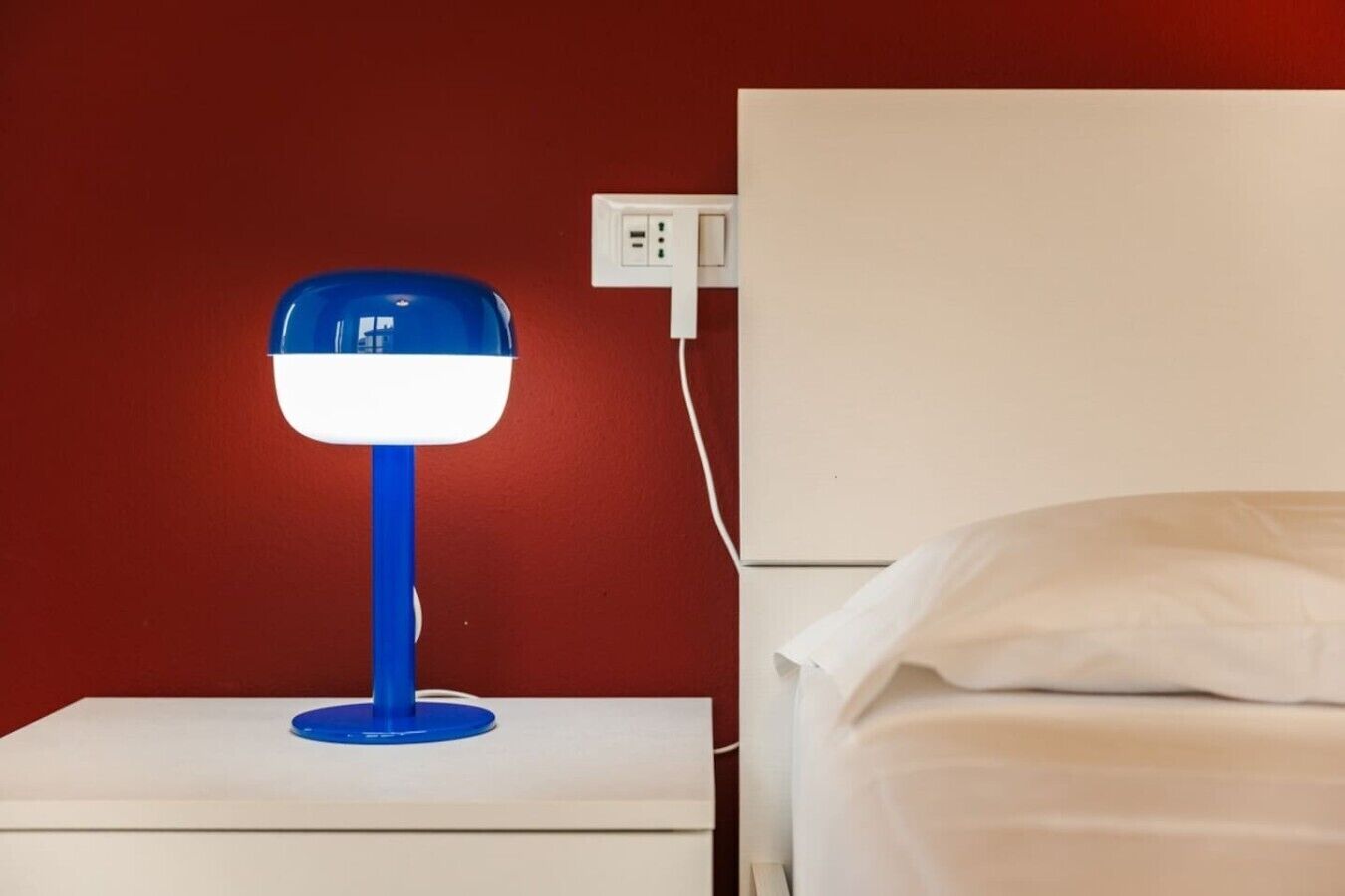 A white bedside table with a blue lamp, a white bed with white bedding, and a red wall.