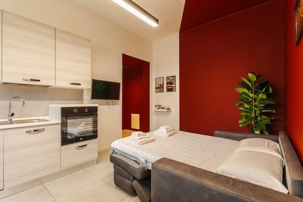 Ein modernes Hotelzimmer mit Bett, Fernseher, Küchenzeile und einer roten Akzentwand.
