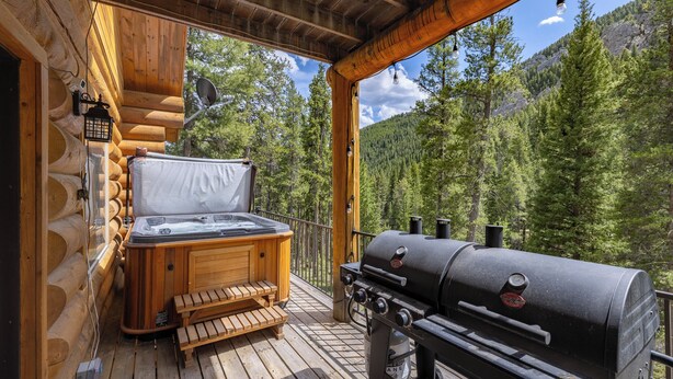 Une terrasse en bois dotée d’un jacuzzi et d’un barbecue, le tout entouré d’une forêt et avec une vue sur les montagnes.