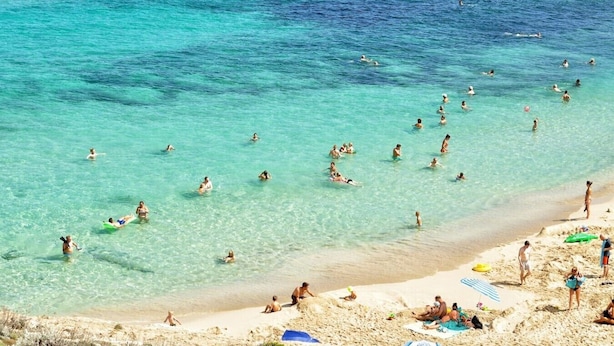 Uma praia com águas turquesa cristalinas, pessoas nadando e tomando sol, e uma orla arenosa com guarda-sóis e cadeiras de praia.