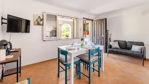 Uma área de refeições com uma mesa e cadeiras, uma televisão presa na parede e um sofá ao fundo.