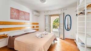 Quarto com cabeceira de madeira, cama com manta bege, ar condicionado branco, porta de vidro deslizante para uma varanda e um armário branco.