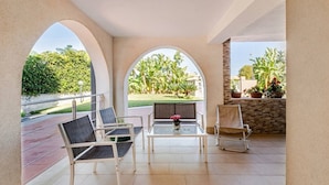 Uma área de estar coberta ao ar livre, com mesa e cadeiras, vista para um jardim paisagístico e um muro de pedra com plantas em vasos.