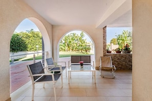 A covered outdoor seating area with a table and chairs, a view of a landscaped garden, and a stone wall with potted plants. - Family Vacation in a Villa with Bikes and a Terrace (Ognina)