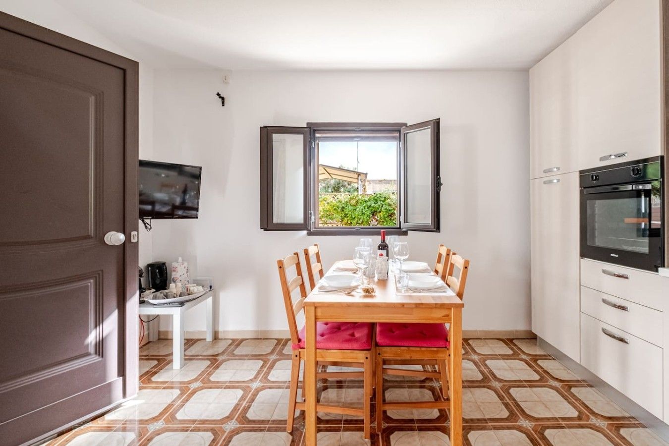 A compact kitchen with a dining table set for four, a built-in oven, and a window with an open shutter.