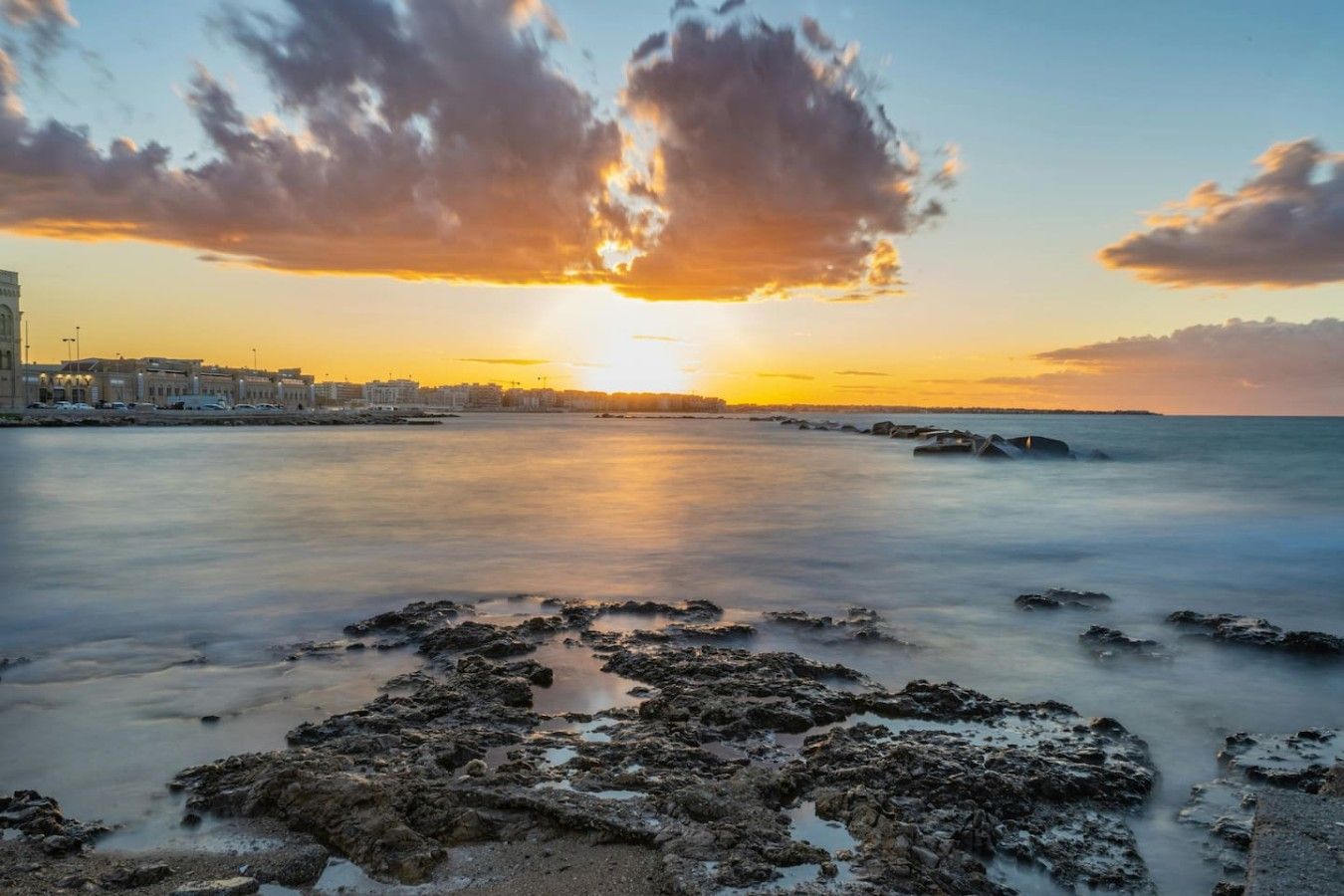 A coastal scene with a sunset, rocky shoreline, and buildings in the background.