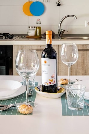A bottle of Merlot wine, two empty wine glasses, and a plate with a pastry on a table in a kitchen setting. - Studio in a Villa · Private Veranda · 200m from the Sea (Ognina)