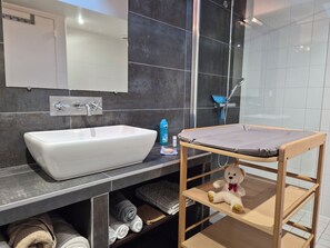 A bathroom with a white sink, dark tiled walls, a glass shower enclosure, and a wooden shelf with towels and a teddy bear. - Villa Locquénolé overlooking Morlaix Bay (Locquénolé)