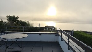 Een terras op het dak, met een tafel, stoelen, een reling en uitzicht op de ondergaande zon over het water.