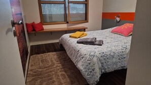 Een slaapkamer met een bed, een bankje, een raam met zonwering, een wandlamp en een vloerkleed.