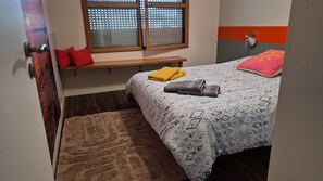 Ein Schlafzimmer mit einem Bett, einer Bank, einem Fenster mit Jalousien, einer Wandlampe und einem Teppich.