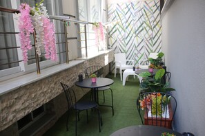 A patio area with a stone wall, a window with pink and white flowers, a metal chair, a round table, and a potted plant.