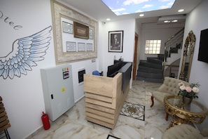 Un espace d’accueil d’hôtel avec un comptoir en bois, des certificats encadrés, une fresque murale, un escalier et une table décorative agrémentée de fleurs.