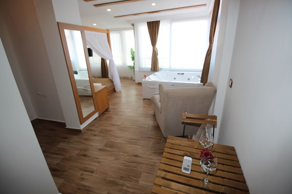 Une chambre d’hôtel avec un plancher en bois, un lit à baldaquin, un coin salon avec une table et une chaise, et une grande fenêtre avec des rideaux.