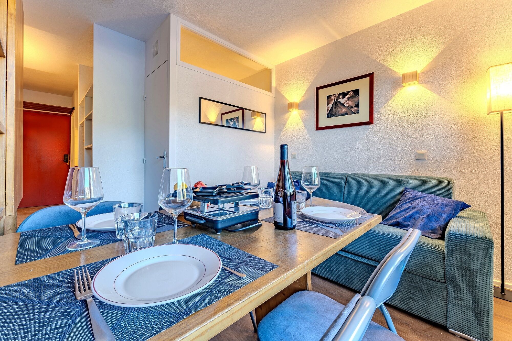 A dining area with a table set for two, a bottle of wine, and a sofa in the background.