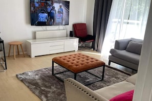 Ein Wohnzimmer mit Sofa, Hocker, Fernseher, rotem Sessel und einem weißen Schrank.