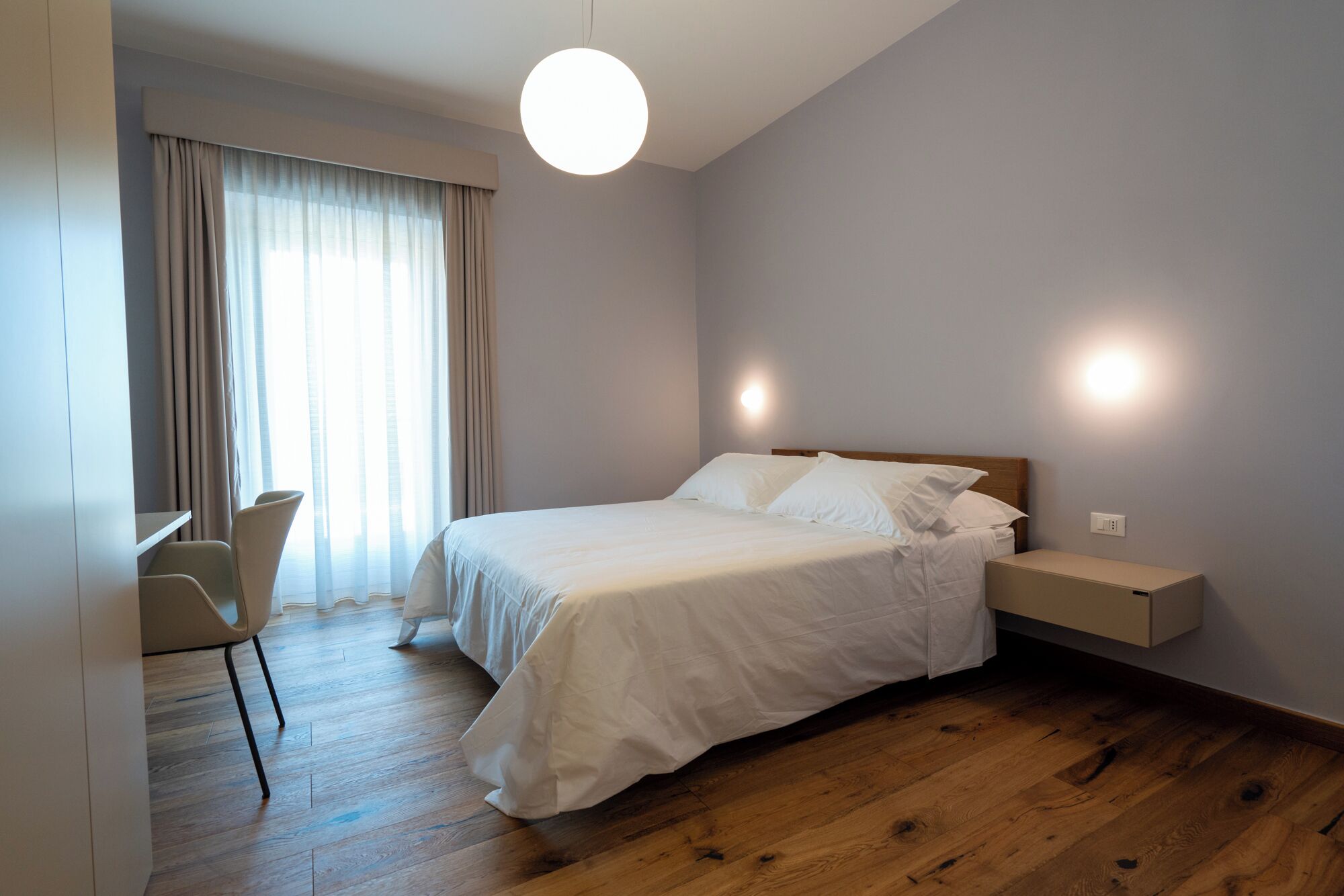 A bedroom with a bed, bedside table, chair, and a large window with curtains.
