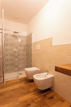 Un bany amb dutxa, dos lavabos, un vàter i bidet, sòl de fusta i un mur revestit de rajoles.