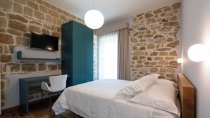 Un dormitorio con pared de piedra, una cama con ropa de cama blanca, un escritorio con una silla y un televisor montado sobre el escritorio.