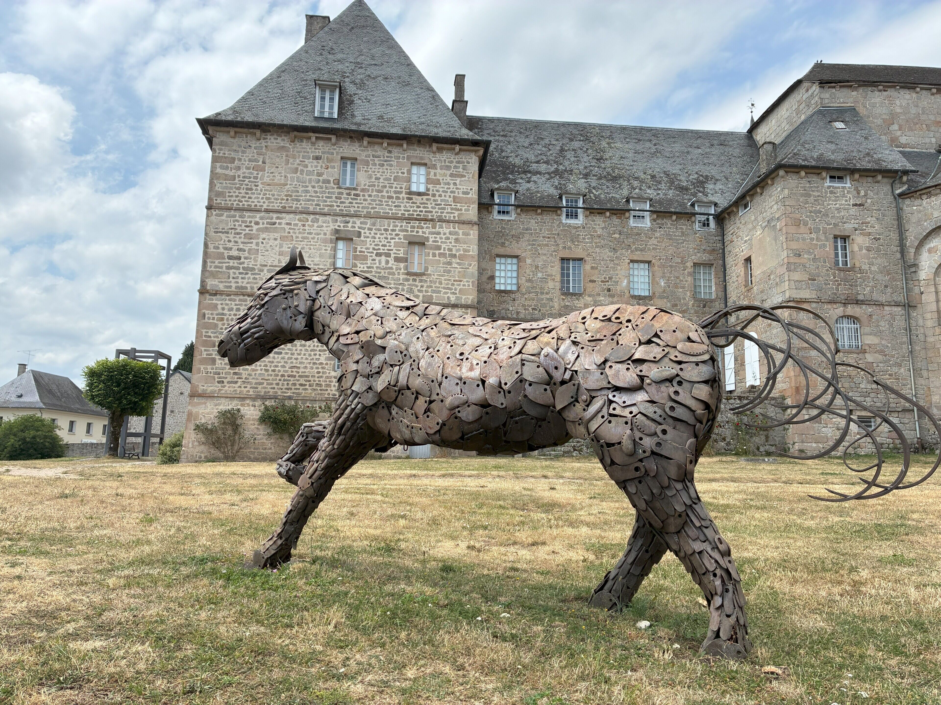 Une sculpture métallique représentant un cheval, située dans un champ verdoyant, avec un bâtiment historique en arrière-plan.