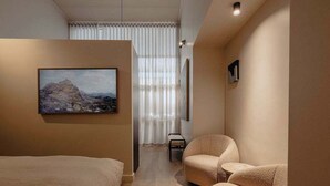 Ett modernt hotellrum med en säng, två fåtöljer, en tavla och ett stort fönster med gardiner.