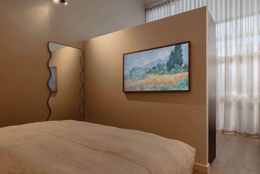 A bedroom with a bed, a painting, a mirror, and a window with curtains.