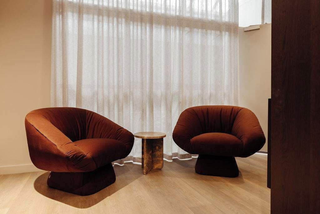 Two brown armchairs, a small round table, and a sheer curtain in a room with wooden flooring.