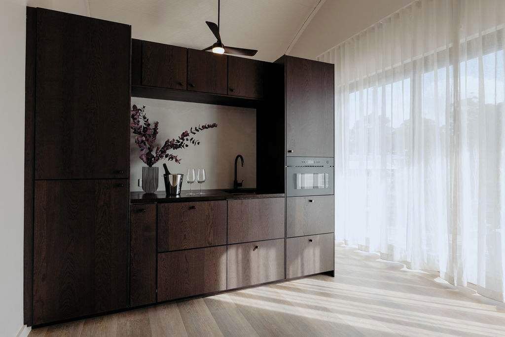 Une cuisine moderne avec des meubles de couleur sombre, un four encastré et une fenêtre ornée de rideaux légers.
