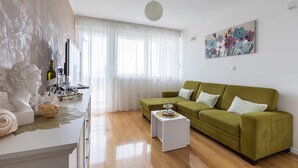 Ein Wohnzimmer mit einem grünen Sofa, einem weißen Couchtisch, einem Gemälde an der Wand und einem Fenster mit transparenten Vorhängen.