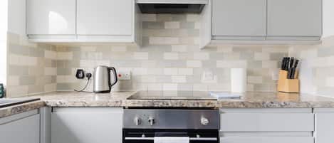 Una cocina con armarios blancos, un hervidor de acero inoxidable, una encimera con un juego de cuchillos y una toalla colgada en la puerta del horno.