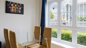 Ein Zimmer mit Glastisch, vier Stühlen, einem Fenster mit Blick auf Gebäude und Grünflächen sowie einem Wandbild.