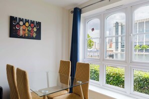 Ein Zimmer mit Glastisch, vier Stühlen, einem Fenster mit Blick auf Gebäude und Grünflächen sowie einem Wandbild.